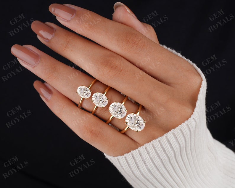 Pode incluir: Cinco alian&ccedil;as de noivado de ouro com diamantes ovais de diferentes tamanhos. As alian&ccedil;as est&atilde;o em uma m&atilde;o que est&aacute; usando um su&eacute;ter branco.