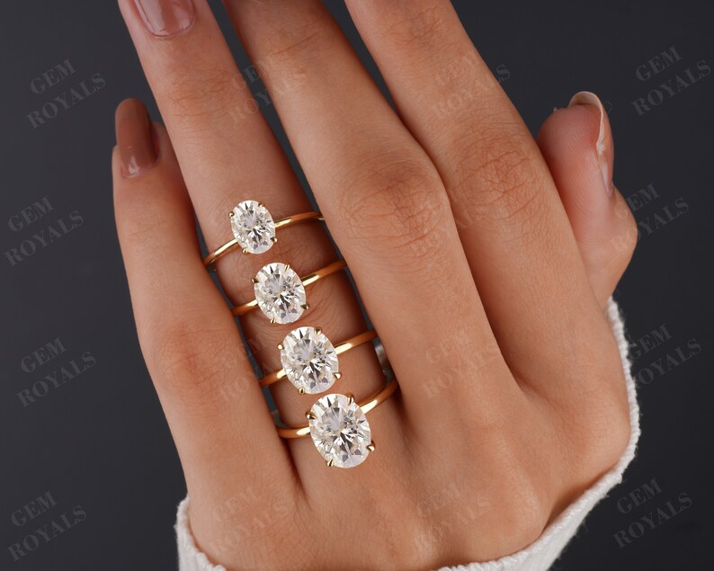 Pode incluir: Cinco an&eacute;is de ouro com diamantes de forma oval de diferentes tamanhos. Os an&eacute;is est&atilde;o empilhados em uma m&atilde;o que est&aacute; usando um su&eacute;ter branco.