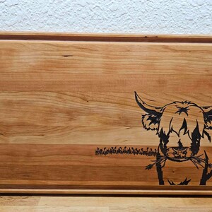 May include: A wooden cutting board with a black silhouette of a Highland cow eating grass. The board has a natural wood grain and a raised edge.