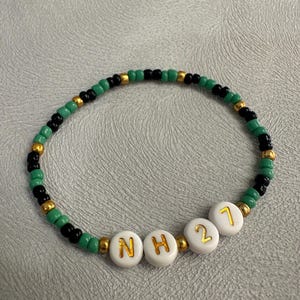 May include: A green, black, and gold beaded bracelet with white letter beads spelling "NH21".