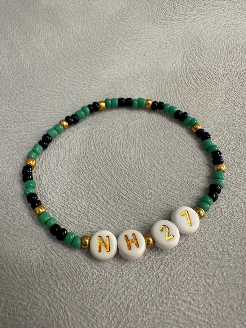 May include: A green, black, and gold beaded bracelet with white letter beads spelling "NH21".
