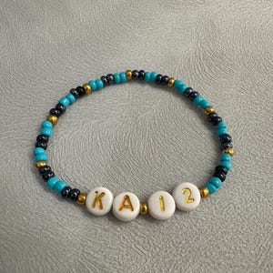 May include: A turquoise and black beaded bracelet with white beads spelling out the name "KAI2".