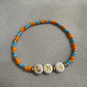 Könnte beinhalten: Ein Armband aus Perlen mit abwechselnd orangefarbenen und blauen Perlen. Das Armband hat weiße Perlen mit goldenen Buchstaben, die "LN4" buchstabieren.
