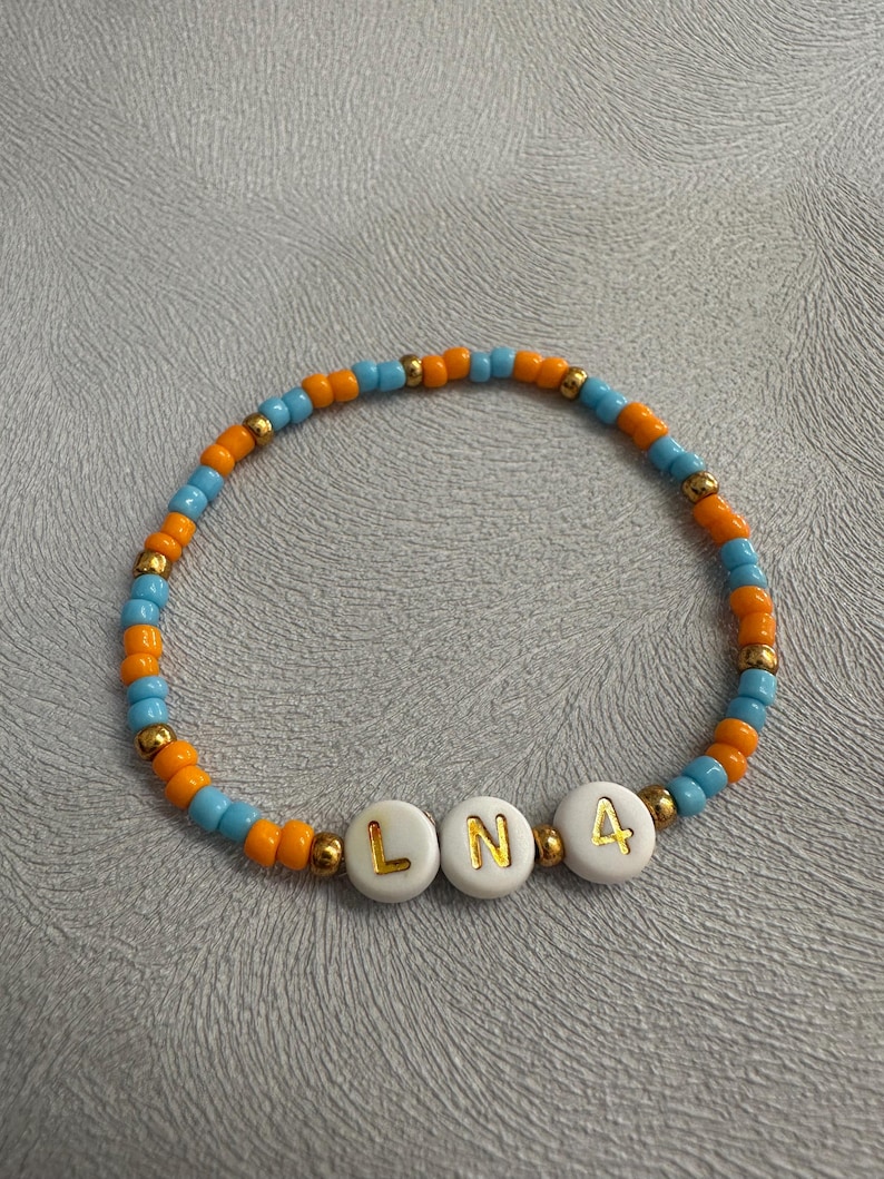 May include: A beaded bracelet with alternating orange and blue beads. The bracelet has white beads with gold lettering spelling out "LN4".