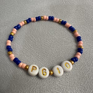 May include: A pink and blue beaded bracelet with gold beads and white letter beads spelling out the name "PGIO".