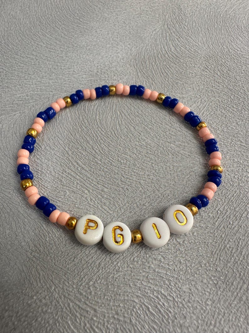 May include: A pink and blue beaded bracelet with gold beads and white letter beads spelling out the name "PGIO".