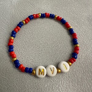 May include: A red, blue, and gold beaded bracelet with white beads that spell out the letters "MVI".