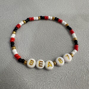 May include: A white beaded bracelet with black and red beads. The bracelet has gold letters spelling out "BEA81".