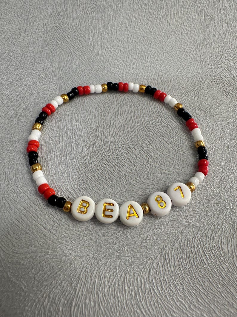 May include: A white beaded bracelet with black and red beads. The bracelet has gold letters spelling out "BEA81".