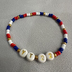 May include: A red, white, and blue beaded bracelet with gold beads and white letter beads spelling out "YT22".