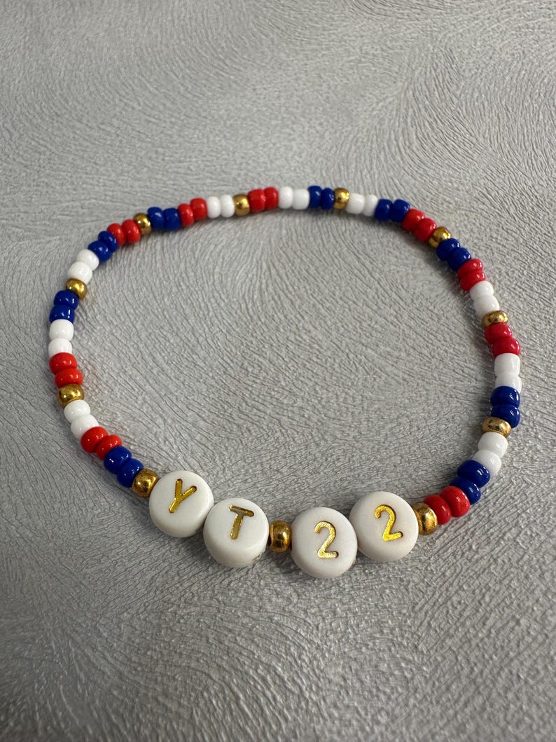 May include: A red, white, and blue beaded bracelet with gold beads and white letter beads spelling out "YT22".