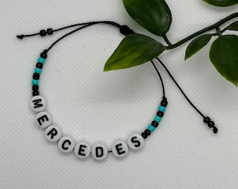 May include: A black string bracelet with white letter beads spelling out the word "MERCEDES" and blue and black spacer beads.
