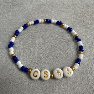 May include: A white beaded bracelet with blue and gold beads. The bracelet has white beads with gold letters spelling out "CS 5"