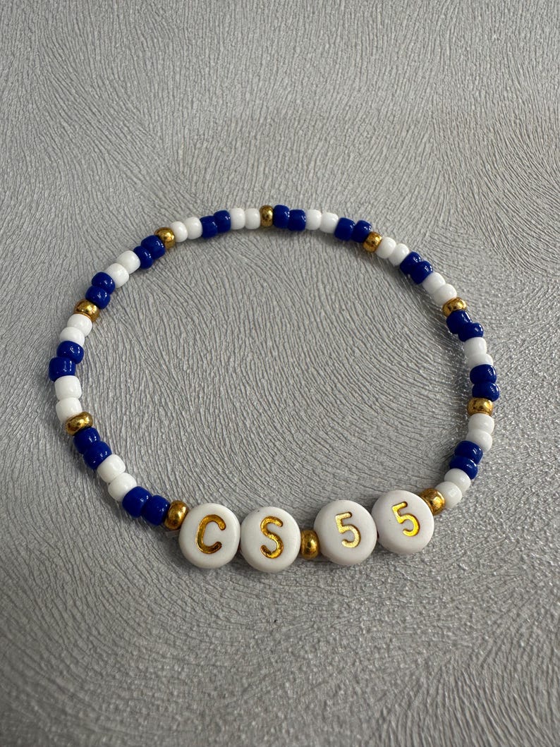 May include: A white beaded bracelet with blue and gold beads. The bracelet has white beads with gold letters spelling out "CS 5"