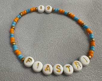 May include: A beaded bracelet with the name "PIASTRI" spelled out in white beads with gold lettering. The bracelet is made with alternating orange and blue beads.