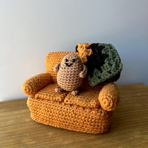 May include: A crocheted brown potato character sitting on a crocheted orange couch with a green and black crocheted blanket.