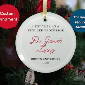 May include: A white ceramic custom ornament with the text "FIRST YEAR AS A TENURED PROFESSOR" and "Dr. Janet Lopez BROWN UNIVERSITY 2024". The ornament is hanging on a Christmas tree.
