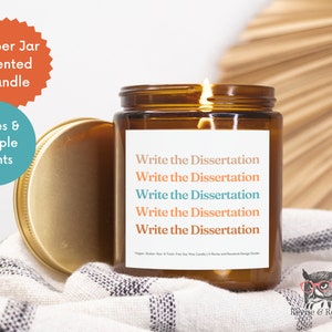 May include: Amber glass jar candle with a gold lid. The candle is labeled "Write the Dissertation" five times. The candle is sitting on a white and black patterned fabric.