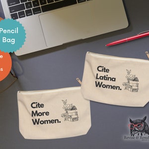 May include: Two canvas pencil bags with gold zippers. One bag reads "Cite More Women." The other reads "Cite Latina Women." Both bags feature a stack of books with floral accents. A laptop and red pen are in the background.