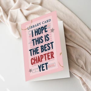 以下が含まれることがあります： 「I HOPE THIS IS THE BEST CHAPTER YET」とネイビーと赤で書かれたピンク色のグリーティングカード。 カードには星のアクセントが付いた図書館カードのデザインがあり、ニュートラルな背景に配置されています。
