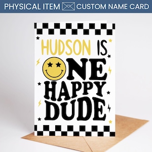 May include: A custom name card with the text "HUDSON IS ONE HAPPY DUDE" in black and yellow. The card features a smiley face with star eyes and lightning bolt accents. The top and bottom borders have a black and white checkered pattern. The card is on a brown envelope.