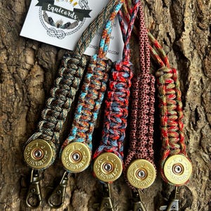 May include: Five lanyards made of braided paracord in various multi-coloured patterns, each featuring a brass 12 gauge E.J. Churchill shotgun shell casing base and a bronze-toned metal lobster clasp. A white business card with the text Equicords is visible.