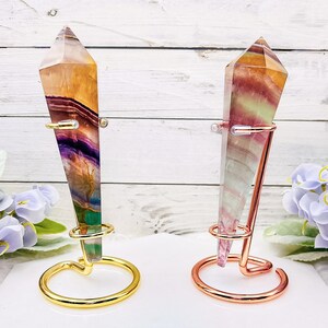 May include: Two rainbow fluorite crystal points on gold and rose gold metal stands. The crystals are iridescent and have a variety of colors, including purple, green, yellow, and pink.