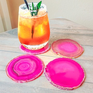 May include: A refreshing drink in a clear glass with a black straw and green garnish, resting on a pink agate coaster. Three additional pink agate coasters are arranged nearby, showcasing natural patterns and vibrant colors. The coasters are approximately 4 inches in diameter.