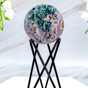 May include: A decorative sphere with a mix of teal, purple, and beige colors, displayed on a black metal stand. The sphere has a textured, organic appearance. The stand has a geometric design.