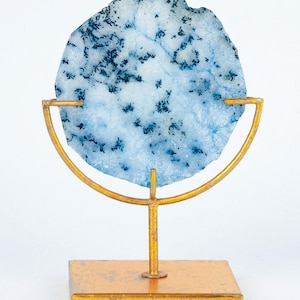 May include: A decorative agate slice in shades of blue and white, mounted on a gold-colored metal stand. The agate features dark blue, tree-like patterns. The stand has a square base and a curved top, creating a modern display piece.