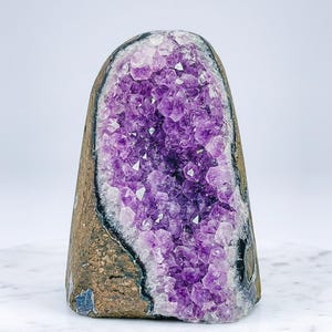 May include: A large amethyst geode with a rough, brown exterior and a cavity filled with sparkling purple crystals. The geode is displayed on a white surface, showcasing its natural beauty and unique crystalline structure.