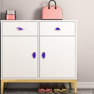May include: A white cabinet with gold legs and purple agate-style drawer pulls. A pink handbag and a small pink and silver box sit on top. Below, several pairs of shoes are visible.