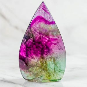 May include: A teardrop-shaped, polished gemstone with a gradient of colors. The stone transitions from a vibrant magenta at the top to a mix of green and yellow at the base. The translucent material has internal patterns.