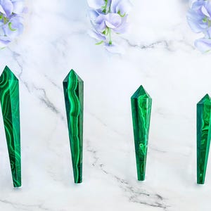 May include: Four green malachite crystal points of varying sizes are arranged on a white marble surface. The crystals have a polished finish, showcasing their natural patterns. Soft purple flowers are in the background.