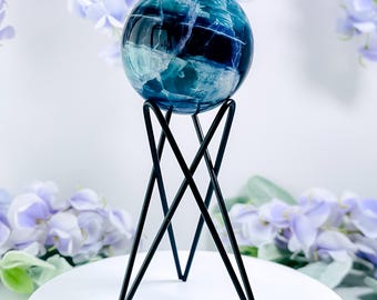 May include: A blue and white crystal sphere sits atop a black metal stand with a geometric design. The sphere has a marbled appearance with shades of blue and white. The stand has a tripod base and a minimalist aesthetic. The background features soft purple flowers.