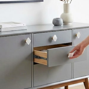 May include: A gray cabinet with diamond-shaped white drawer pulls. The cabinet has three drawers and two doors. The drawer is being opened, revealing a wooden interior. The cabinet sits on wooden legs.