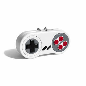 May include: A white pendant in the shape of a retro video game controller, with a silver chain. It has a grey directional pad and a grey section with four red buttons. The design is inspired by classic gaming consoles.