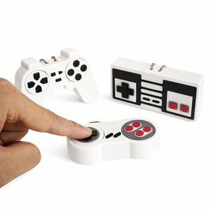 Puede incluir: Tres mini mandos de juego blancos con detalles en negro y rojo. Un dedo presiona un mando. Los mandos están diseñados para parecerse a los mandos de videojuegos clásicos.