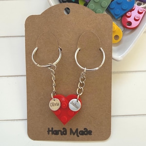 Può includere: Due portachiavi con un ciondolo a forma di cuore rosso realizzato con mattoncini ad incastro. Ogni portachiavi ha un anello e una catena argentati, con un piccolo tag rotondo inciso con un nome. Le parole "Hand Made" sono stampate sulla carta.
