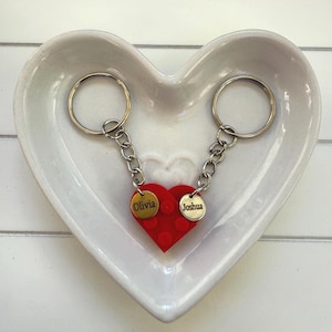 May include: Two keychains connected to a red heart-shaped charm with the names "Olivia" and "Joshua" on gold-colored tags. The keychains are in a heart-shaped white ceramic dish.