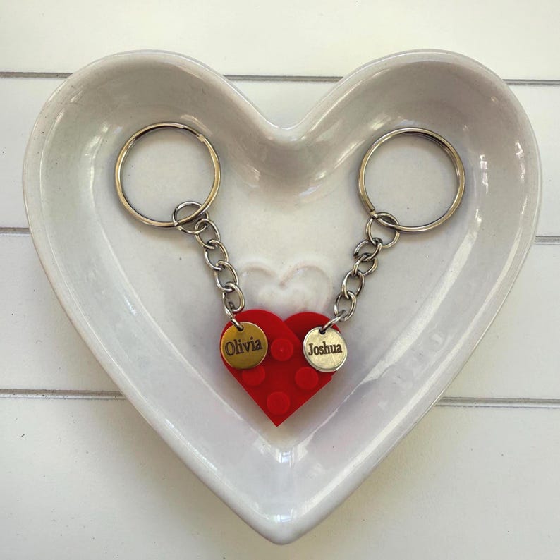 May include: Two keychains connected to a red heart-shaped charm with the names "Olivia" and "Joshua" on gold-colored tags. The keychains are in a heart-shaped white ceramic dish.