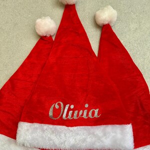 May include: Three red felt Santa hats with white fur trim. The middle hat has the name "Olivia" embroidered in silver glitter.