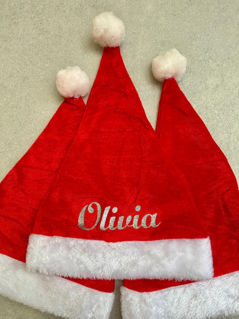 May include: Three red felt Santa hats with white fur trim. The middle hat has the name "Olivia" embroidered in silver glitter.