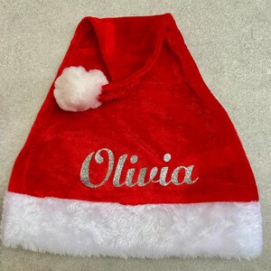 May include: Red velvet Santa hat with a white faux fur trim and a white pom-pom. The hat has the name "Olivia" written in silver glitter.