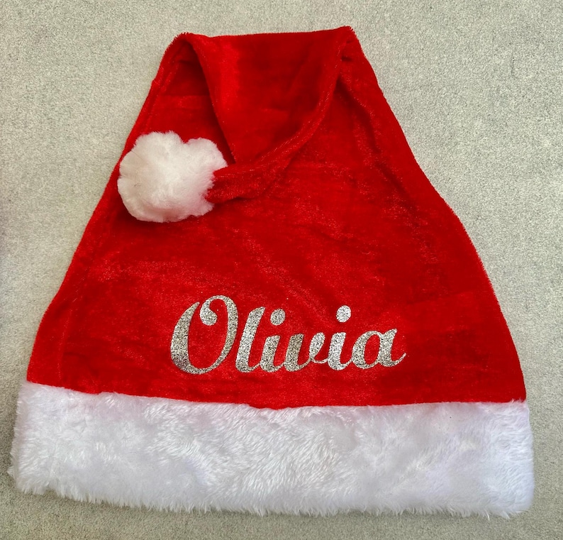 May include: Red velvet Santa hat with a white faux fur trim and a white pom-pom. The hat has the name "Olivia" written in silver glitter.