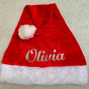 May include: Red velvet Santa hat with a white faux fur trim and a white pom-pom on top. The hat has the name "Olivia" written in silver glitter.