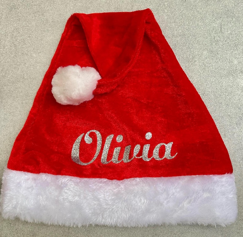 May include: Red velvet Santa hat with a white faux fur trim and a white pom-pom on top. The hat has the name "Olivia" written in silver glitter.