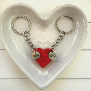 May include: A red heart-shaped keychain with the names "Joshua" and "Olivia" engraved on separate silver tags. The keychain is connected to two silver keyrings by a chain. The items are displayed in a white heart-shaped dish.