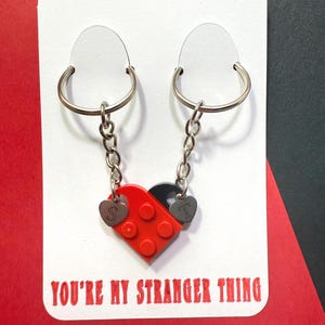 Può includere: Due portachiavi con un ciondolo a forma di cuore rosso e nero realizzato con blocchi ad incastro. Ogni portachiavi ha un anello e una catena color argento. Il ciondolo ha le lettere "S" e "T" su piccole targhette a forma di cuore. La carta recita "YOU'RE MY STRANGER THING."
