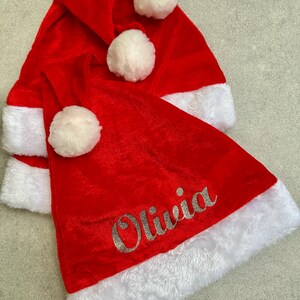 May include: Three red velvet Santa hats with white fur trim and white pom-poms. The bottom hat has the name "Olivia" embroidered in silver glitter.